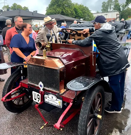 Johanna Olofsson, kommunstyrelsens ordförande i Surahammar, och Västmanlands landshövding Johan Sterte står vid den vinröda Vabisbilen från 1903 under lunchstoppet i Köping vid Styrkeprovet 2025. De samtalar med en förare från Aros Motorveteraner.